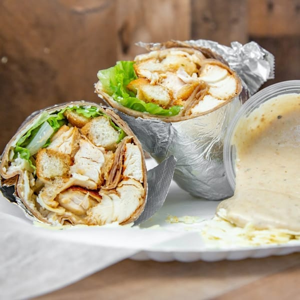Two chicken Caesar wraps with lettuce and croutons, one with dressing spilling from a container.